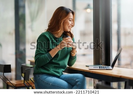 Beautiful young Asian woman relaxing holding coffee and smiling brightly with laptop in cafe.