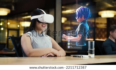 Young asian woman wearing VR headset conversing with a cartoon character avatar via an futuristic screen hologram. Futuristic communication scifi concept. 3D rendering picture. Augmented reality. Royalty-Free Stock Photo #2211712387