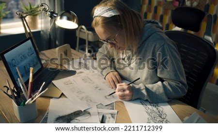 Female animator creates sketches for the cartoon. Looks at the laptop screen infront of her and draws on the paper. Comics Compilation. Storyboard movie layout for pre-production. Royalty-Free Stock Photo #2211712399