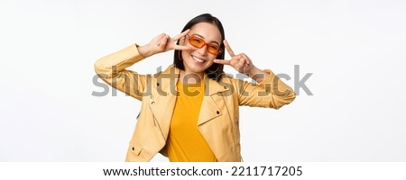 Portrait of stylish asian modern girl, wearing sunglasses and yellow jacket, showing peace, v-sign gesture, standing over white background, happy smiling face