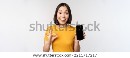 Excited asian girl showing mobile phone screen, okay sign, recommending smartphone app, standing in yellow tshirt over white background