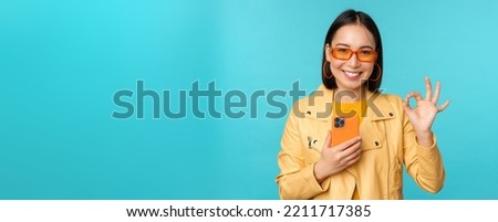 Smiling asian girl with smartphone, shwoing okay, ok sign in approval, standing over blue background