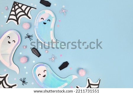 Cute pastel colored Halloween party flat lay with ghost shaped plates, spider web napkins and confetti on blue background
