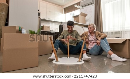 Caucasian girl looking at her middle eastern husband or boyfriend assembling table on floor. Young smiling multiethnic couple moving to their new apartment. Home relocation. Modern spacious flat Royalty-Free Stock Photo #2211747423