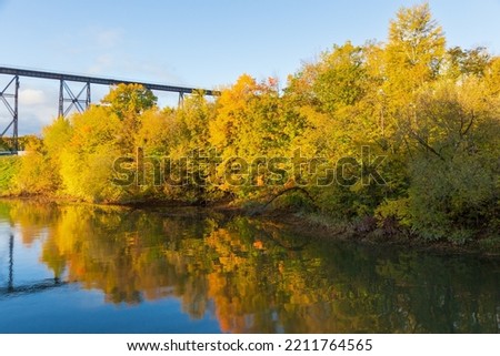 View of yellow foliage and its reflection in the Cap-Rouge River seen during a fall morning golden hour, with the 1908 railway trestle bridge in the background, Quebec City, Quebec, Canada Royalty-Free Stock Photo #2211764565
