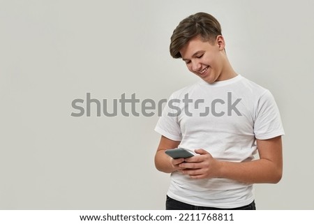 Smiling caucasian teenage boy browsing his smartphone. Guy of zoomer generation wearing t-shirt. Concept of modern youngster lifestyle. Isolated on white background. Studio shoot. Copy space