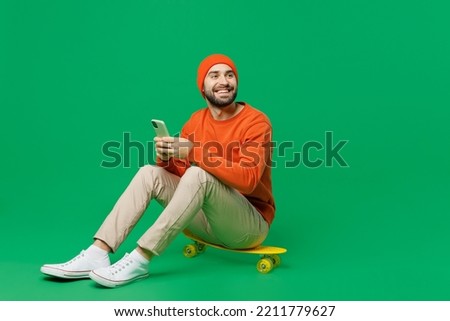 Young happy minded pensive fun cool caucasian man 20s wearing orange sweatshirt hat sit on skateboard hold use mobile cell phone look aside isolated on plain green color background studio portrait.