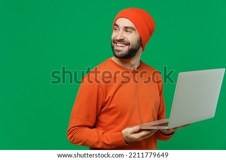 Young smiling happy man 20s in orange sweatshirt hat hold use work on laptop pc computer browsing internet look aside on workspace area copy space isolated on plain green background studio portrait. Royalty-Free Stock Photo #2211779649