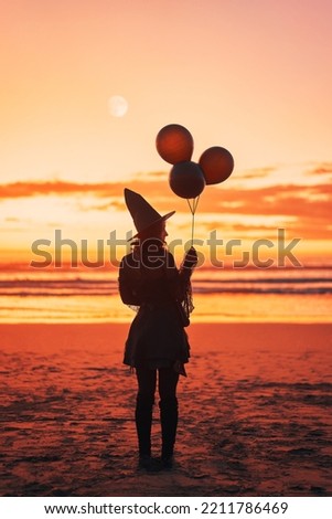silhouette of a woman dressed as a witch with balloons on the shore of the beach at sunset	