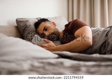 A young man writes text messages on a mobile phone lying in bed in a bedroom at home.