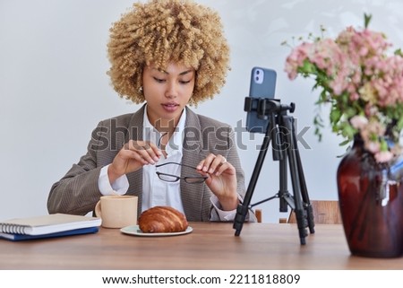Elegant curly female entrepreneur has online meeting with colleagues holds spectacles poses in front of smartphone camera has delicious lunch talks about important issues being in cafeteria.