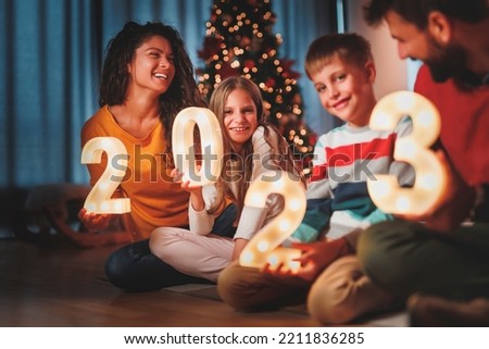 Parents celebrating New Years Eve at home with kids, sitting by the Christmas tree, holding illuminative numbers 2023 representing the upcoming New Year Royalty-Free Stock Photo #2211836285