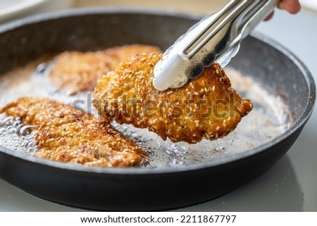 Detail of pliers taking fried schnitzel out of a sizzling pan. Royalty-Free Stock Photo #2211867797
