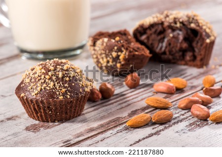 Close-up picture with selective focus only on one cupcake, also cup of milk and four chocolate cupcakes one of them broken decorated with almonds and hazelnut on wooden table in cafe 