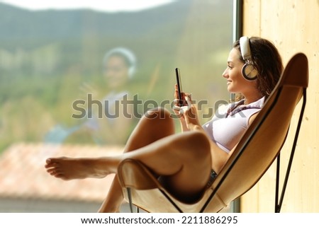 Happy woman at home or hotel listening to music sitting on a chair looking through a window