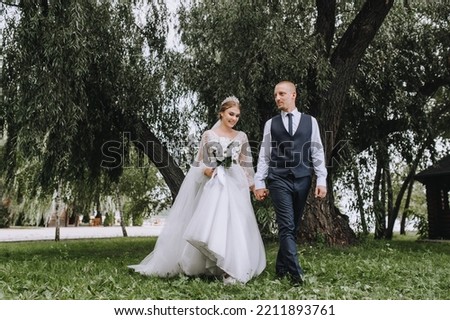 A stylish groom in a blue vest and a beautiful smiling bride are walking in a park in nature on green grass, holding hands. Wedding photography, portrait of newlyweds in love.