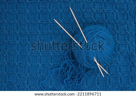 A blue ball of thread for knitting and two knitting needles on a blue knitted fabric. Top view.