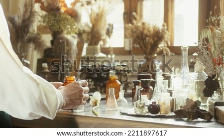 A close-up of a perfumer at his desk looking for a new fragrance. Vintage cinematic perfumery concept. Sunny day in the working workshop. Lots of ingredients, glass flasks. Depth of field Royalty-Free Stock Photo #2211897617