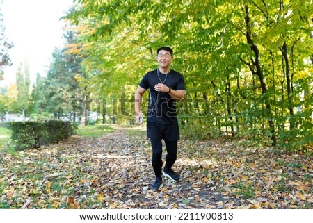 Man on morning jog in autumn park, asian sportsman in sportswear with headphones and phone in forest listens to music on jog and walk active lifestyle.