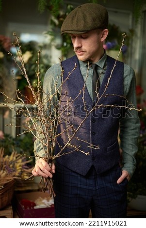 Cute caucasian male in peaked cap and vintage clothes from 20s holding bouquet made of branches standing in flower store. Mother's Day or Valentine's Day concept. High quality vertical image