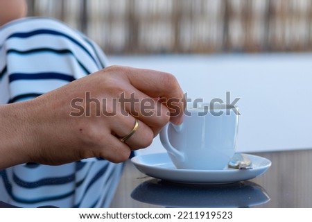 a woman hand with gold rings holds a cup of coffee from a white saucer on a gray glass table in an outdoor terrace