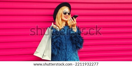 Portrait of stylish young woman with shopping bags and smartphone using voice command recorder, assistant or takes calling in city wearing blue fur coat, black round hat on pink background