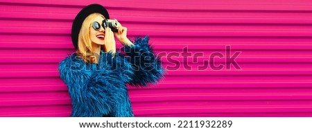 Portrait of stylish happy smiling young woman photographer with film camera taking picture wearing blue fur coat, black round hat on pink background