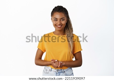 Image of smiling Black woman holding hands together and looking at camera helpful, assisting customer or client, standing friendly against white background Royalty-Free Stock Photo #2211939097