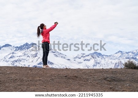 young woman standing in red jacket, taking a picture with her phone in the middle of the Andes Mountains of Chile