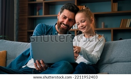 Cheerful father and little beautiful daughter girl sit on couch watch movies cartoons on internet spend time together at home having fun use laptop play games online modern tech usage parental control