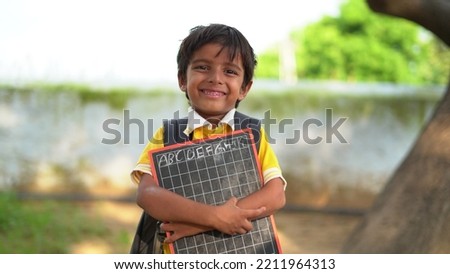 Portrait of happy cute little indian boy in school uniform holding blank slate against orange background, Adorable elementary kid showing black board. child education concept. rural india. Royalty-Free Stock Photo #2211964313