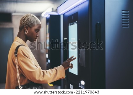A side view of a young beautiful elegant woman with painted white very short hair paying for service underground parking or buying a subway or train ticket using an electronic self-service kiosk Royalty-Free Stock Photo #2211970643