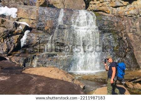 a male Tourist taking pictures on a smartphone Gadelsha waterfall in the Southern Urals on a spring sunny day on the Irendyk ridge