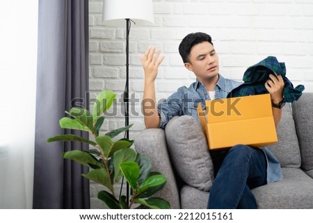 Young Asian man dissatisfied opening parcel at home, sitting on couch with cardboard box, angry displeased customer confused by wrong or damaged order, bad delivery shipping service concept Royalty-Free Stock Photo #2212015161