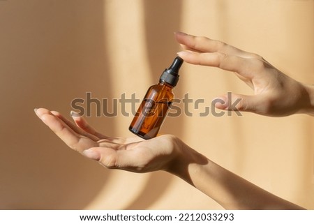 Bottle of serum in women's hands. Glass bottle with dropper cap in women's hands. Amber glass container with dropper lid for cosmetic products on brown background in sunlight. Royalty-Free Stock Photo #2212033293
