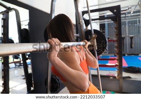 Young and beautiful woman with a sculpted body doing squat exercises with a dumbbell bar on her neck. The woman is wearing orange. Concept Gymnastics, health and wellness.