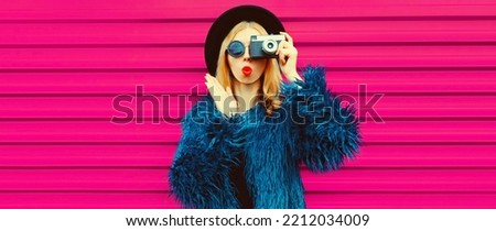 Portrait of stylish blonde young woman photographer with film camera taking picture and blowing her lips sends air kiss wearing blue fur coat, black round hat on pink background