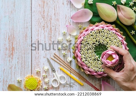 Pink lotus petal krathong for Thailand Loy Krathong festival decorates from lotus, crown flower, incense stick and candle with blurred focus of hand holding lotus flower on wooden background.