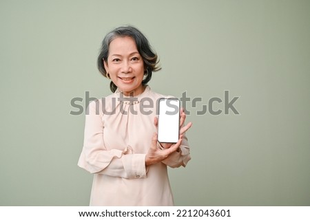 Attractive 60s senior asian woman in casual clothes holding or showing a smartphone white screen mockup, standing isolated over green studio background. online deal, mobile application