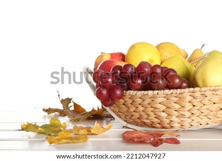 Wicker bowl with fresh fruits and autumn leaves on table against white background, closeup. Harvest festival