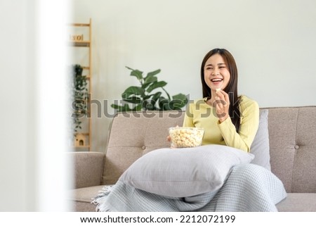 Young asian woman putting a bowl of popcorn on pillow and eating popcorn while sitting on the big comfortable sofa to watching movie on television in living room at home. Royalty-Free Stock Photo #2212072199