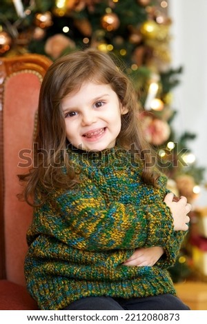 A little girl dressed in a green knitted sweater sits in a room with a Christmas decoration.