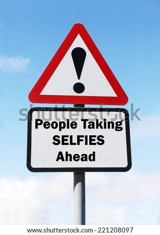 A red and white triangular road sign with a warning about people taking selfies concept against a partly cloudy sky.