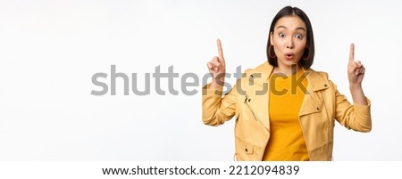 Surprised asian girl pointing fingers up, express interest, showing advertisement ahead, demonstrating promo banner on top, standing against white background