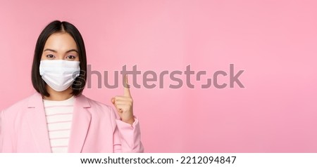Close up portrait of asian businesswoman in medical face mask and suit, pointing finger up, showing advertisement, top banner, standing over pink background