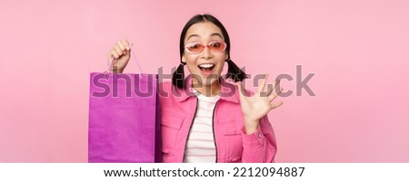 Shopping. Stylish asian girl in sunglasses, showing bag from shop and smiling, recommending sale promo in store, standing over pink background