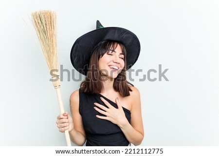 Young caucasian woman dressed as a witch holding a broom isolated on blue background laughs out loudly keeping hand on chest.