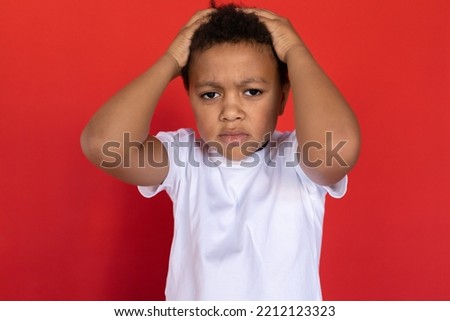 Portrait of desperate preteen boy holding head in hands. Upset multiracial child wearing white T-shirt standing with frustrated expression against red background. Stress and depression concept