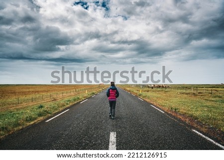 One female tourist running on straight road through farmland and stormy sky in countryside on summer at Iceland