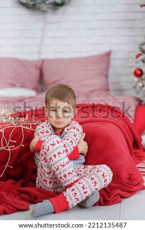 The boy is sitting near the bed and the Christmas tree with a red plaid and a checkered bedding in New Year's pajamas in a lotus position with an agitated expression on his face. Holidays Eve, Christm